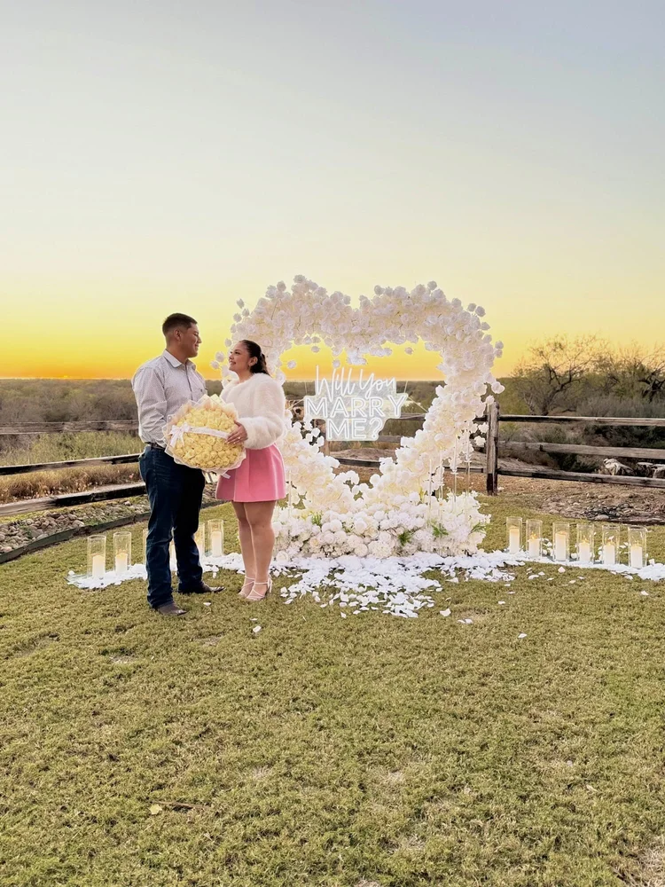 Couple celebrating their engagement in front of a white rose heart arch with a Will You Marry Me neon sign, candles and white petals at sunset