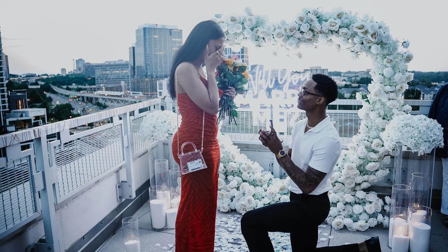 A man proposes marriage to a woman on a rooftop decorated with white roses and candles, with city buildings in the background.