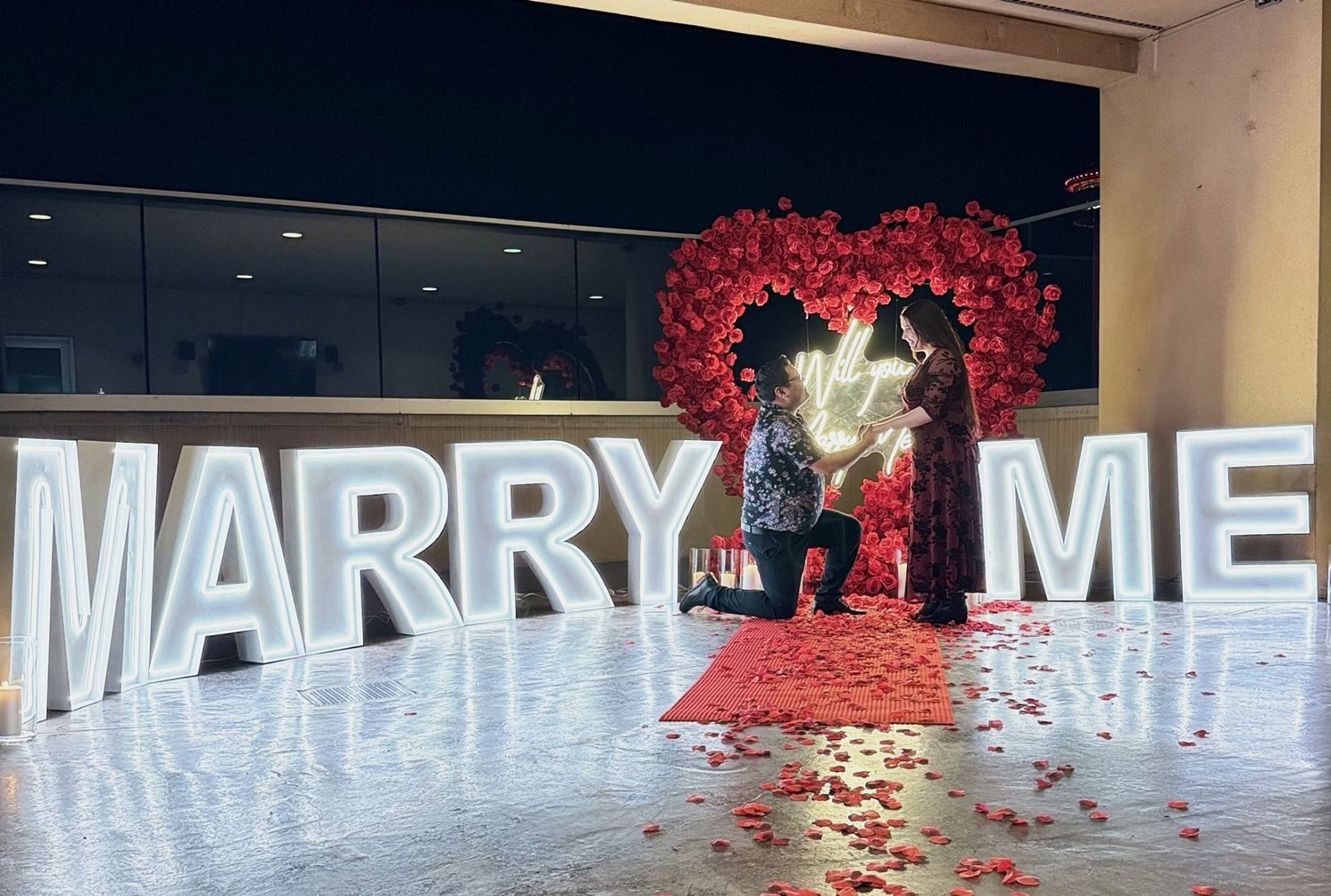 Man proposing on one knee in front of a red rose heart arch with a neon sign and large lit MARRY ME letters at night