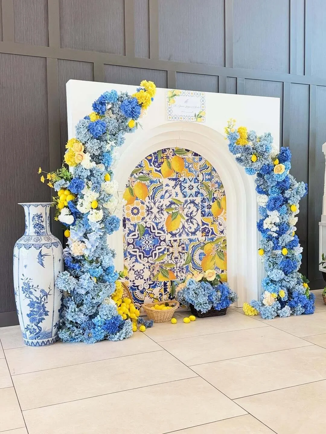 A decorative floral setup with blue and yellow flowers, a large white vase with blue floral patterns, a tiled backdrop with intricate blue, yellow, and white patterns, and a white archway, on a beige tiled floor.