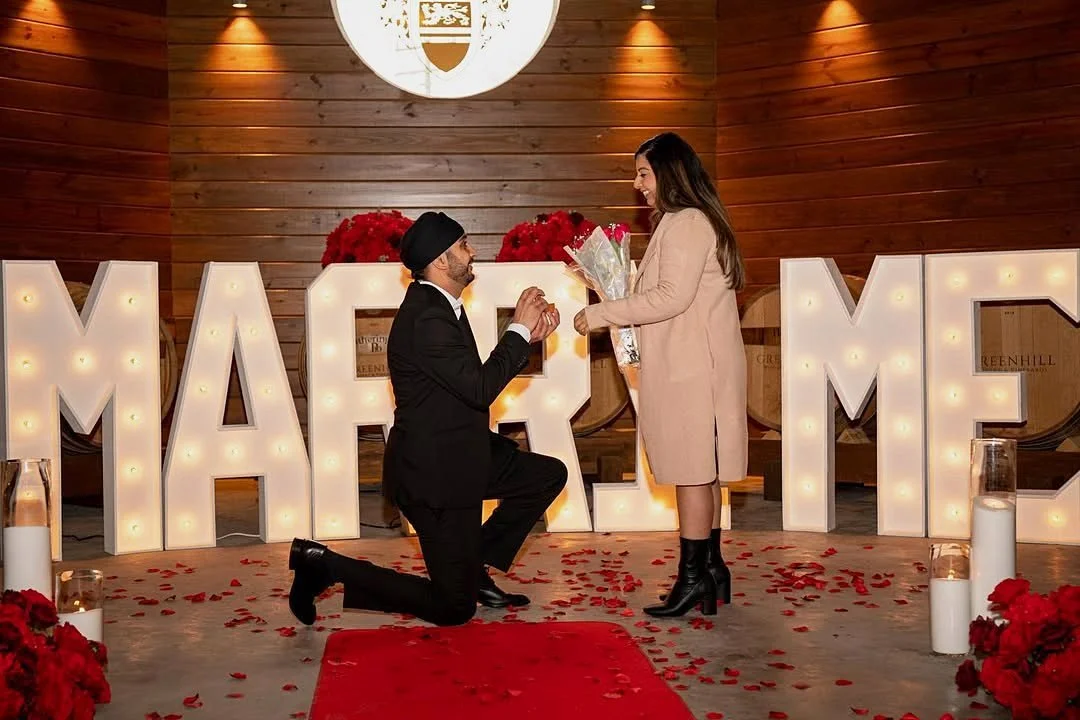 Man proposing on one knee in front of lit MARRY ME marquee letters on a red carpet with rose petals and candles at Greenhill Vineyards in Middleburg, Virginia