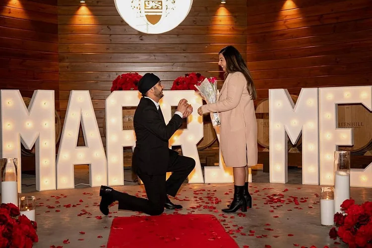 Man proposing on one knee in front of lit MARRY ME marquee letters on a red carpet with rose petals and candles at Greenhill Vineyards in Middleburg, Virginia