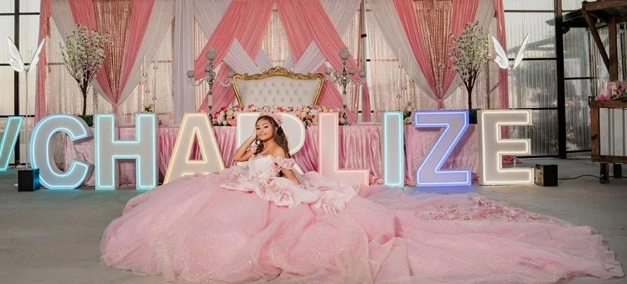 Quinceañera honoree in a pink ball gown seated in front of rainbow lit marquee name letters, a pink and white draped backdrop and a gold throne chair