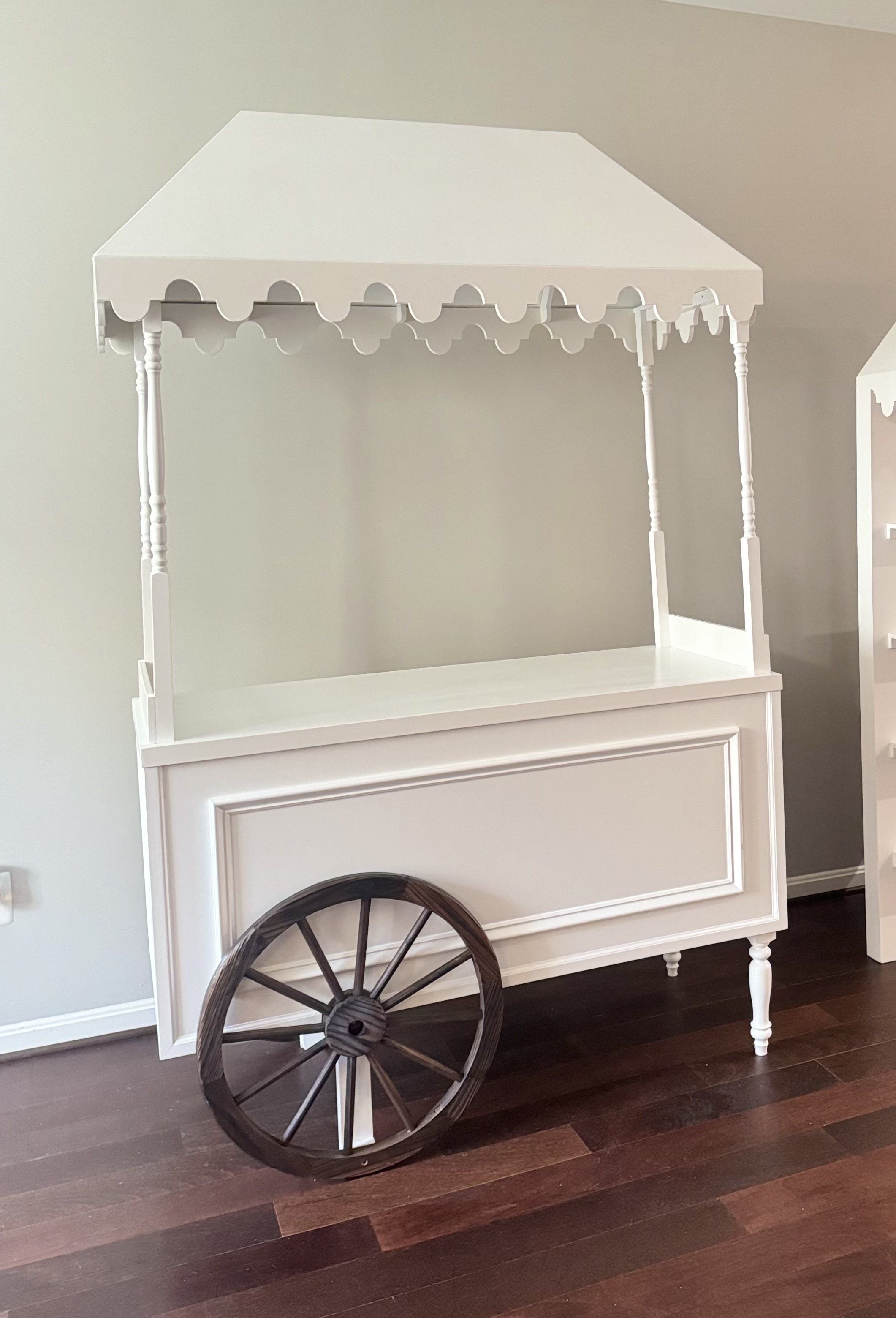 A white wooden cart with a canopy, decorative trim, and turned legs, placed on a dark wood floor.