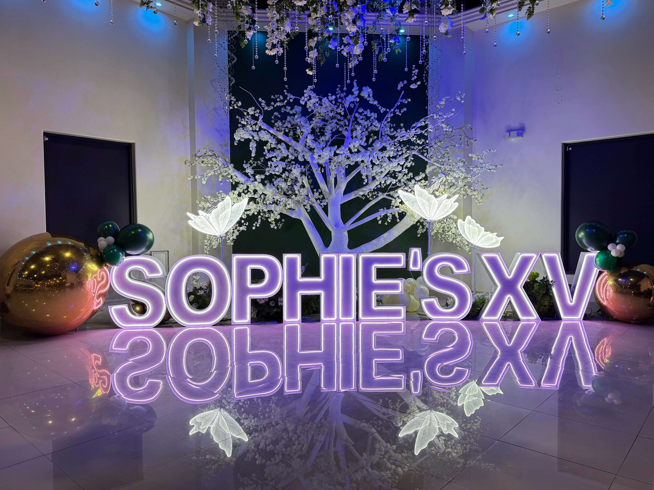 Decorative event display featuring a large illuminated sign that reads 'SOPHIE'S XV', reflections on the shiny floor, a white floral tree in the background, and gold and metallic balloon arrangements with smaller balloons, set against a neutral colored wall.