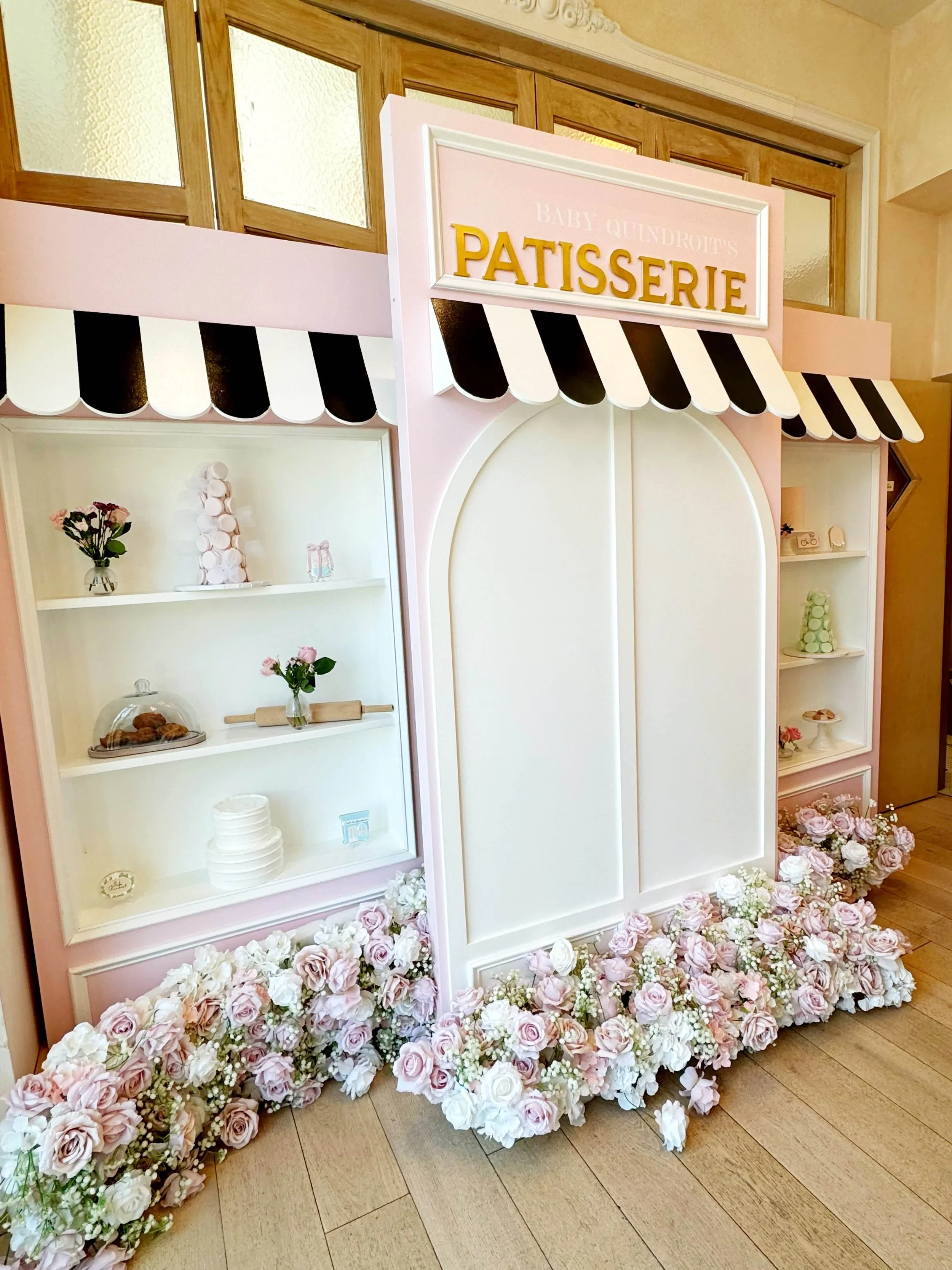 Pink and white bakery display with flowers at the base, a sign reading 'Patisserie' and shelves with pastries and decor