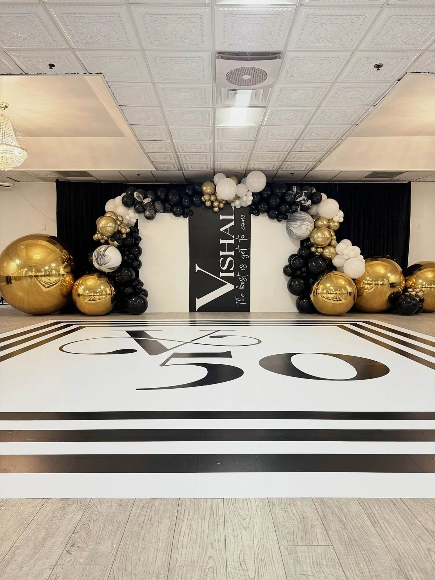 50th birthday setup with a black, white and gold balloon garland, mirrored gold spheres, a custom backdrop and a printed dance floor wrap