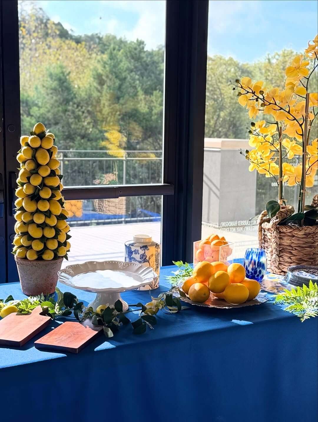 Table decorated with yellow lemons and yellow orchids near a window with a view of trees.