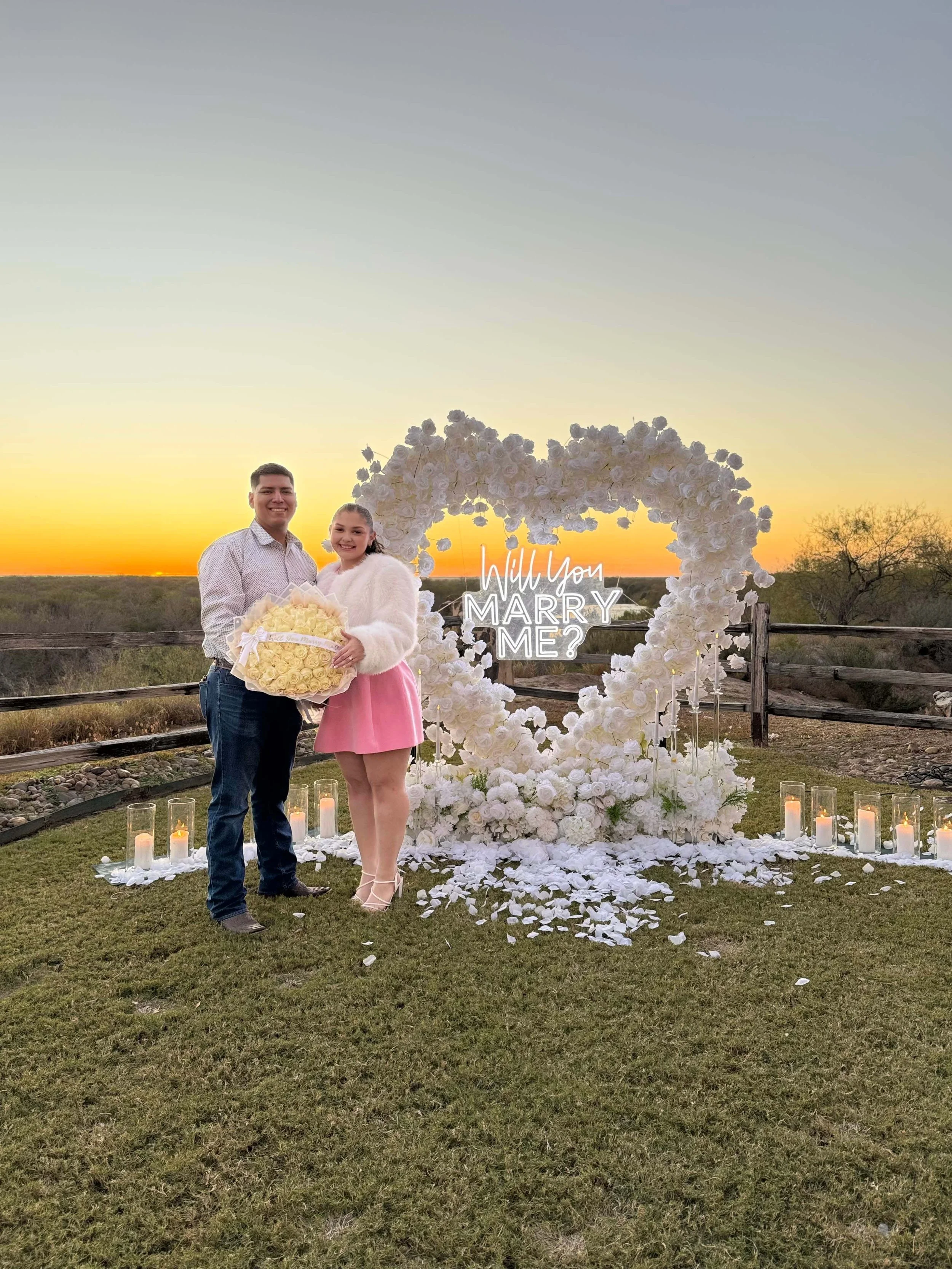 A couple standing outdoors at sunset in front of a large heart-shaped floral arrangement with a sign that says 'Will You Marry Me?' The man is holding a bouquet of yellow roses, and they are smiling. Candles are placed along the ground behind them.