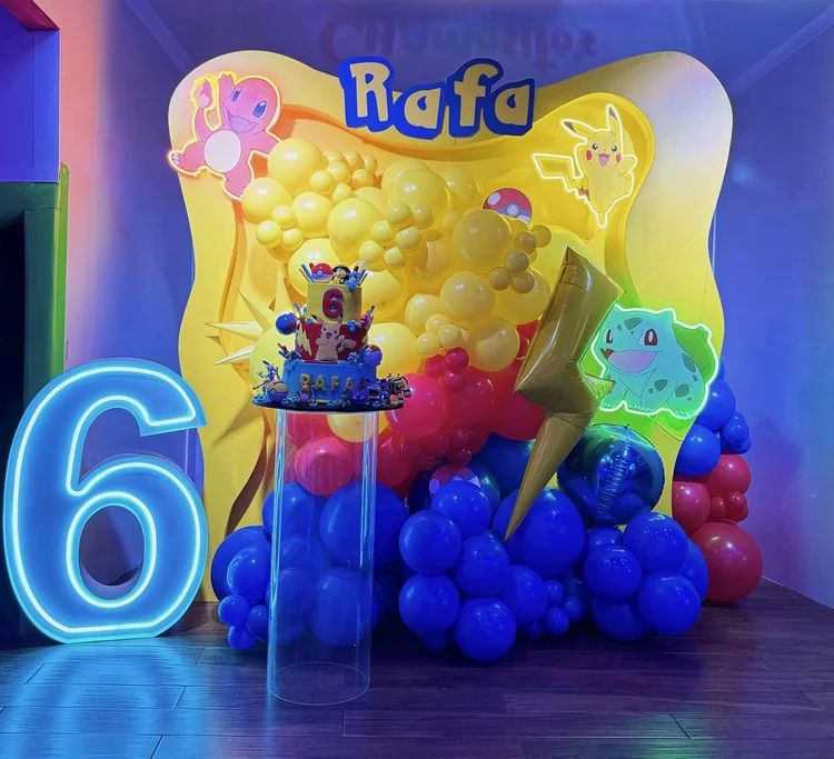 Pokémon themed sixth birthday setup with a yellow backdrop, neon character signs, balloon garlands, a lit blue neon number 6 and an acrylic cake stand
