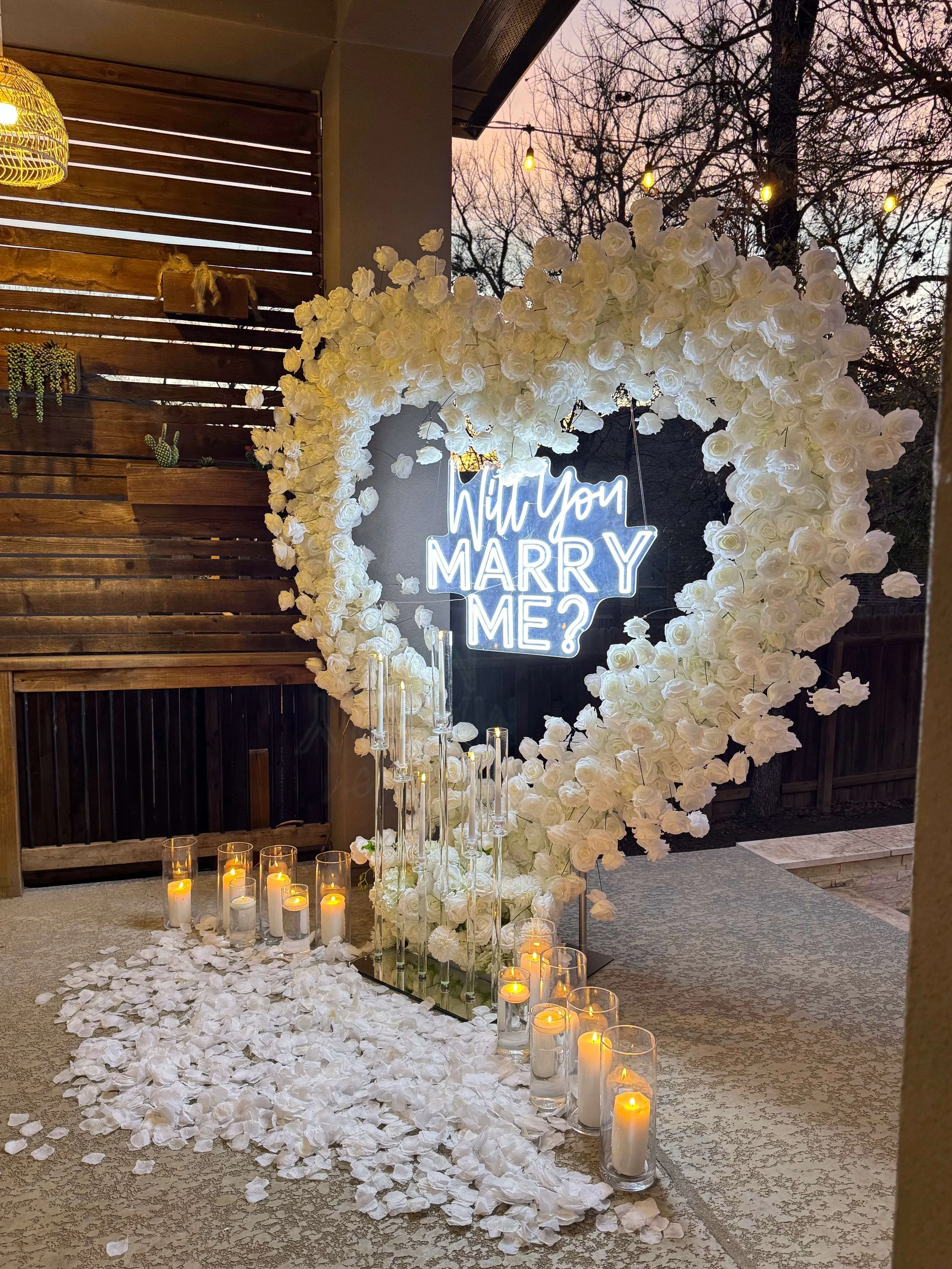 A large white floral heart display with a neon sign asking 'Will you marry me?' surrounded by candles and flower petals, set outdoors at sunset.