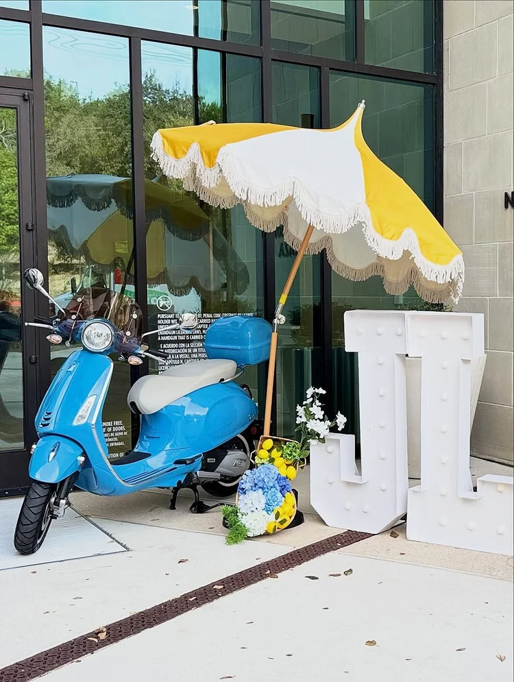 Italian lemon themed party entrance with a blue scooter, a yellow and white fringed umbrella, lit white marquee letters, lemons and blue hydrangeas
