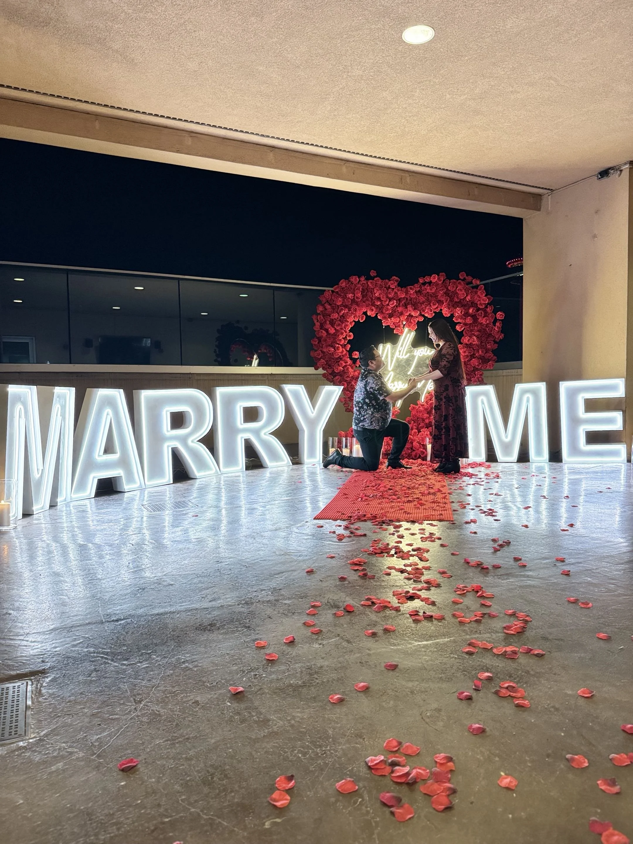 A couple shares a romantic moment during a wedding proposal. The man is kneeling on one knee, holding the woman's hand. They are standing in front of a decorative backdrop with a heart made of pink roses and illuminated words that say 'Will you marry me?'. Rose petals are scattered on the floor, and large illuminated letters spelling 'MARRY ME' are in the foreground.