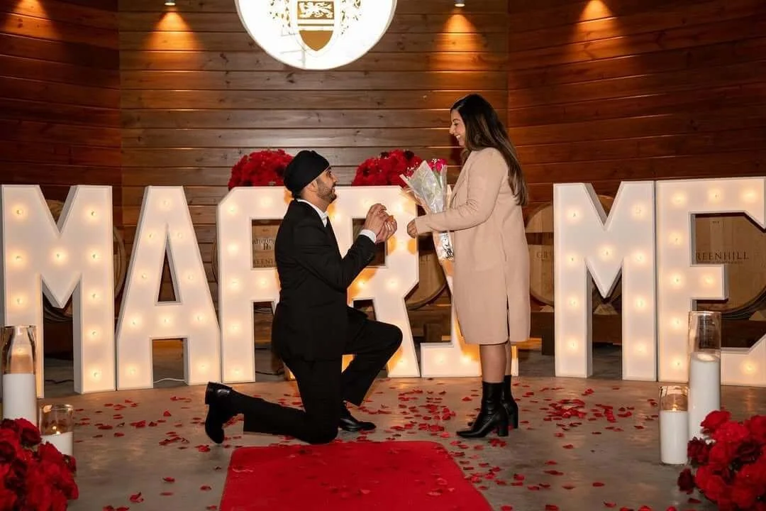 A man proposing to a woman in front of large illuminated letters spelling 'MARRY ME' with rose petals on the floor and a woman holding a bouquet of flowers.