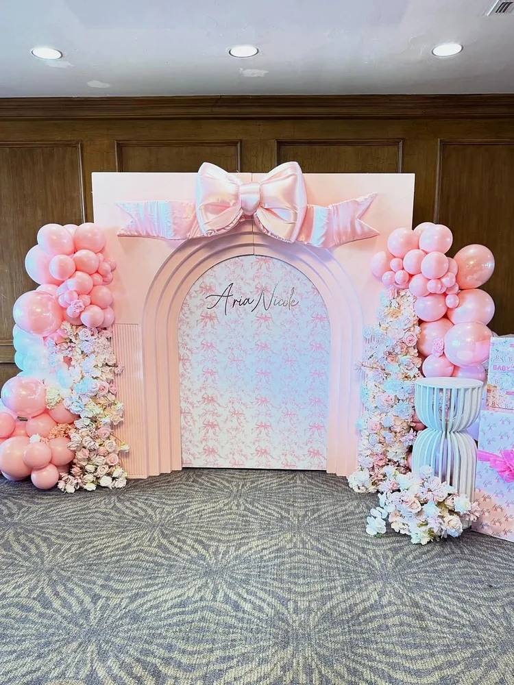 Baby shower backdrop with a pink arch topped with a satin bow, pink balloon garlands, blush floral arrangements, a white pedestal and a custom name sign