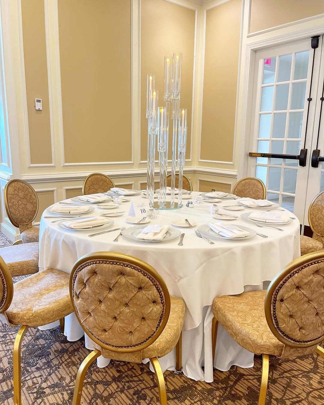 Round banquet table set with white tablecloth, plates, napkins, cutlery, and glasses, with a tall glass centerpiece in a decorated room with beige walls and chairs with gold frames and patterned upholstery.