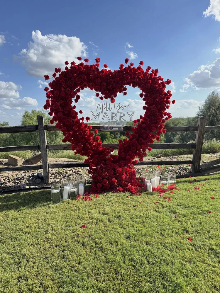 Outdoor proposal setup with a red rose heart arch, Will You Marry Me neon sign, glass candle holders and scattered rose petals on a lawn