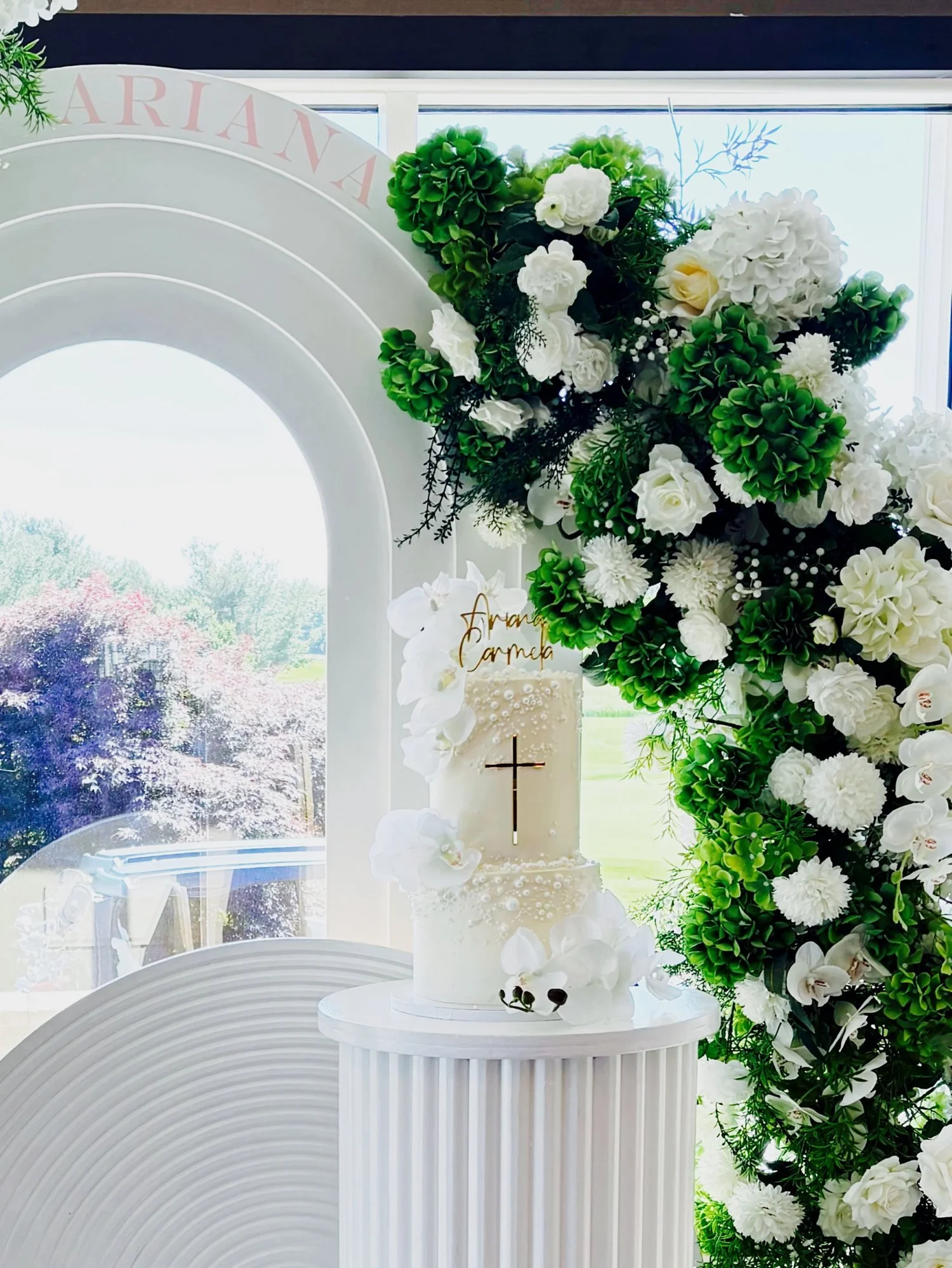 Christening cake display on a white fluted pedestal beside an arch backdrop and a white and green hydrangea floral arch at Chantilly National Golf & Country Club in Centreville, Virginia