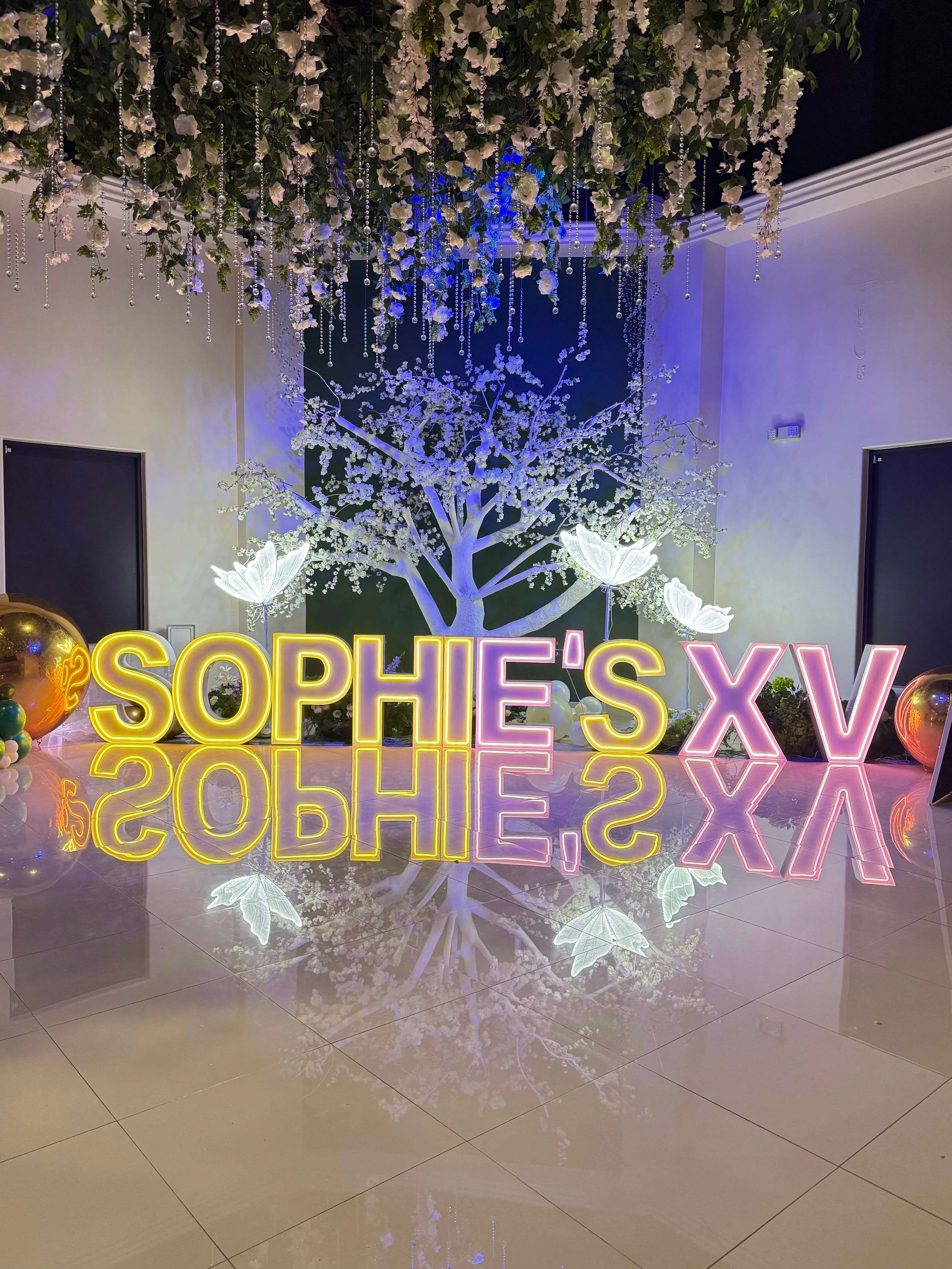 Neon sign reading 'SOPHE'S XV' in yellow and pink letters, reflected on the shiny floor, with white illuminated floral and tree decorations in the background, including hanging white flowers and butterflies