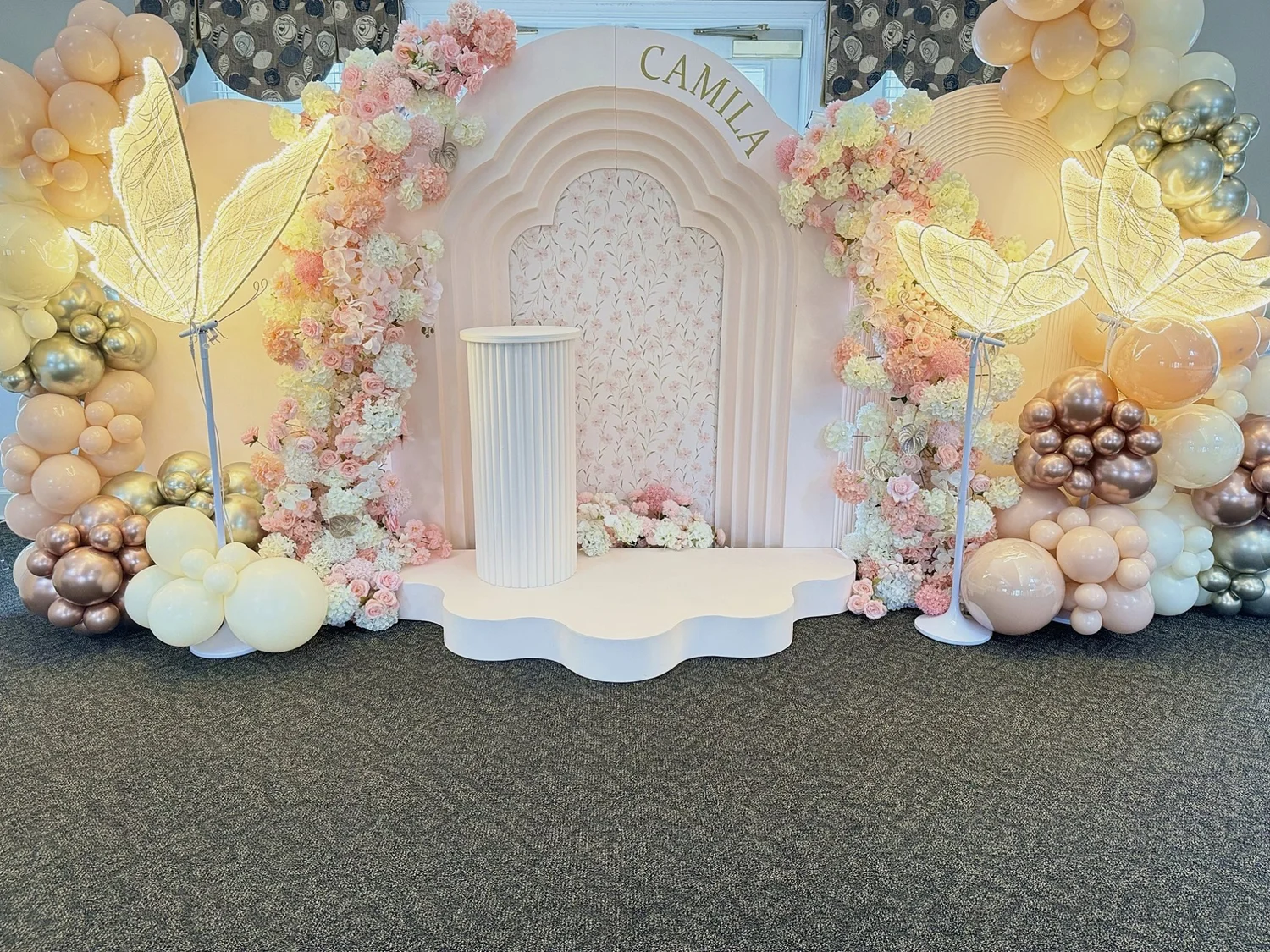 Butterfly themed birthday setup with a tiered arch backdrop, pink and cream floral arches, light-up butterflies, a scalloped stage, fluted pedestal and peach, cream and rose gold balloon garlands in Fredericksburg, Virginia