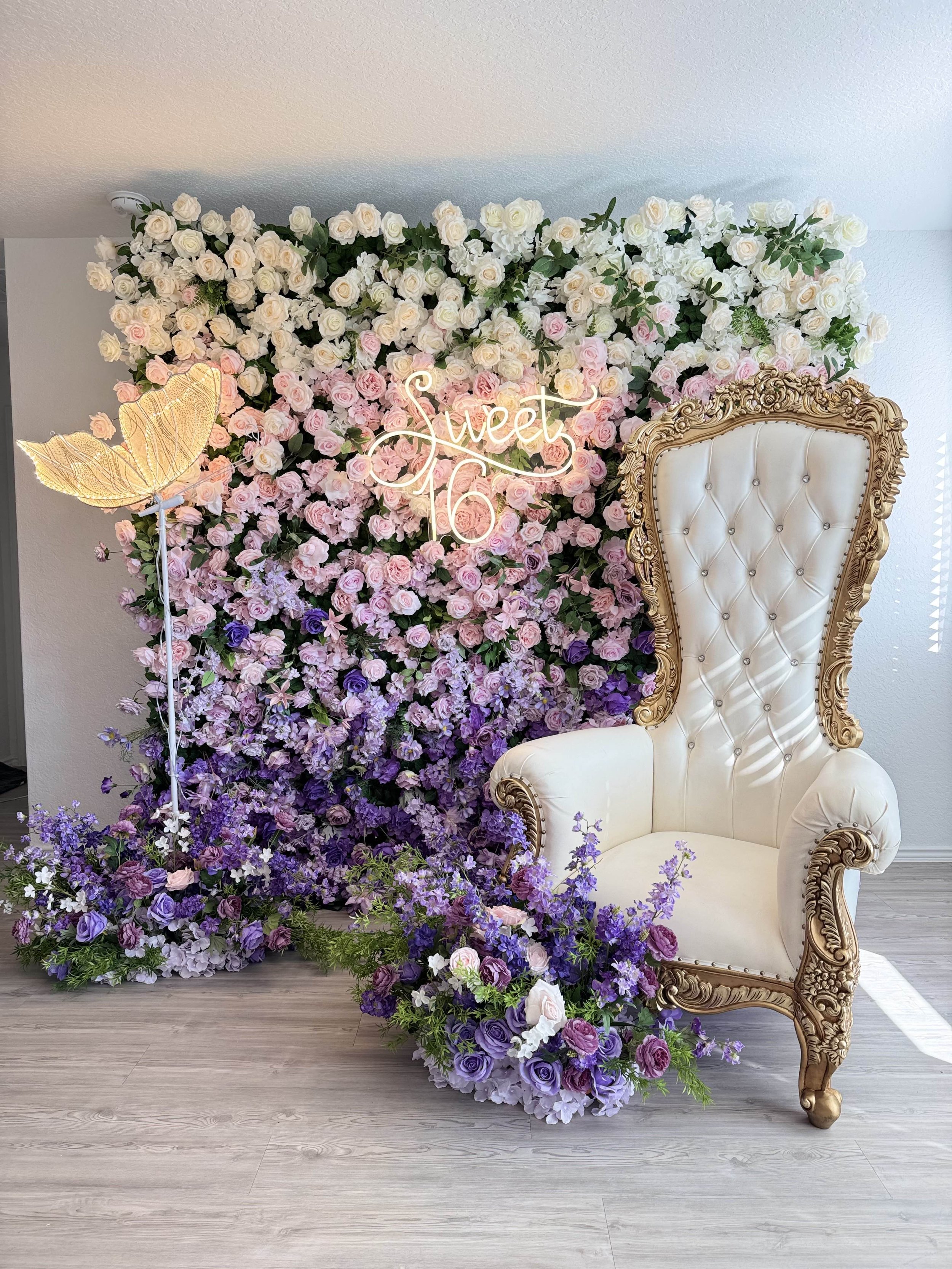 Floral backdrop with pink, white, and purple flowers, a neon sign that says 'Sweet 16', a white and gold ornate high-back chair, and purple flower arrangements on the floor.
