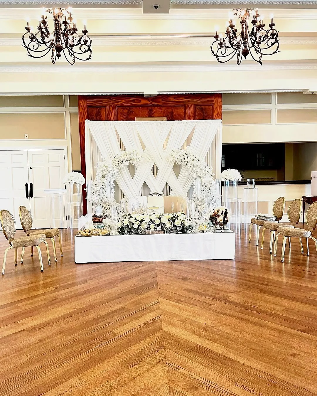 Engagement party sweetheart table on a mirrored stage with a white draped crisscross backdrop, white floral arches and clear acrylic pedestals at the Country Club of Fairfax in Virginia