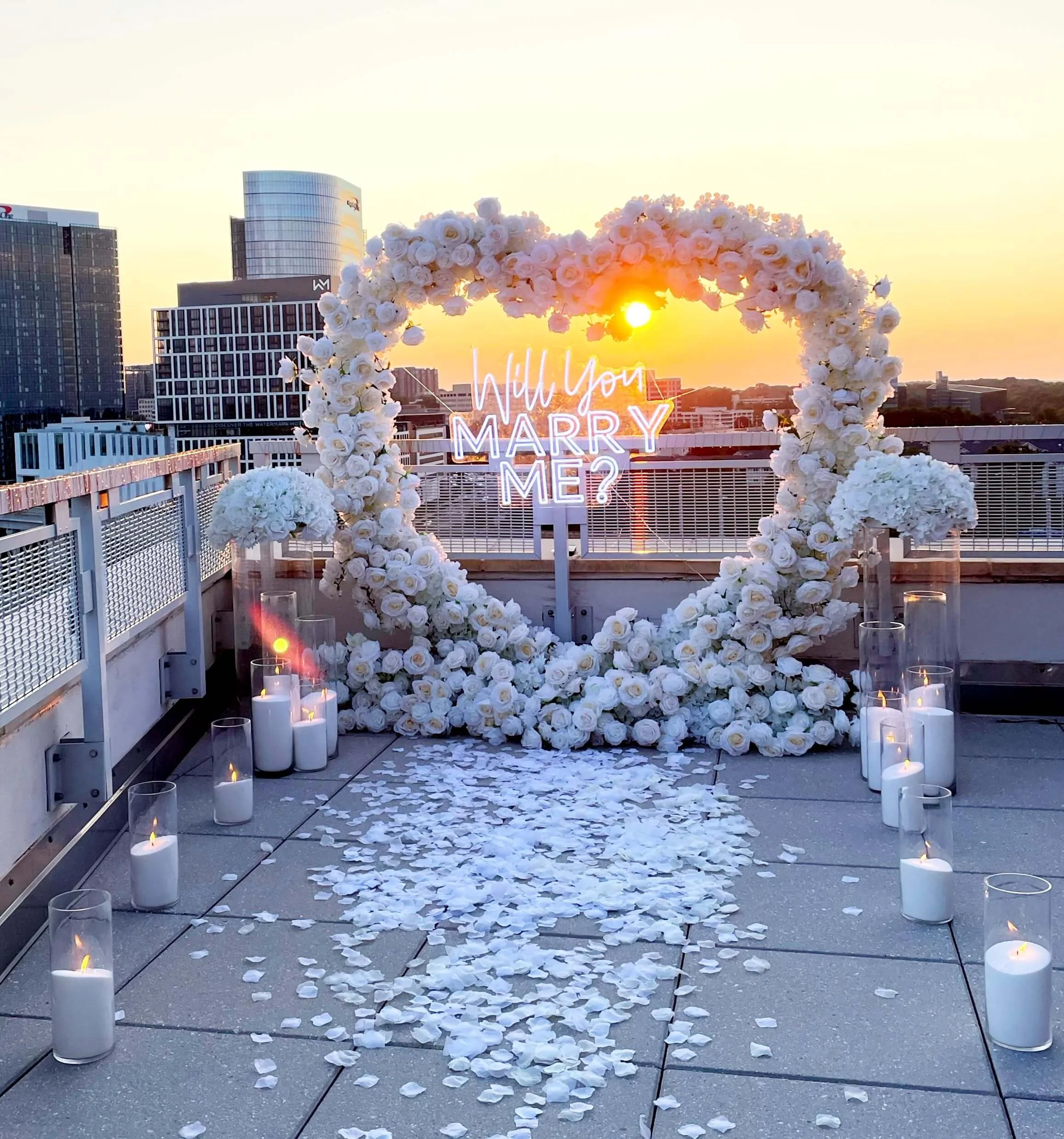 A rooftop wedding setup with a heart-shaped floral arch, candles, and a neon sign that says 'Will You Marry Me?' during sunset.