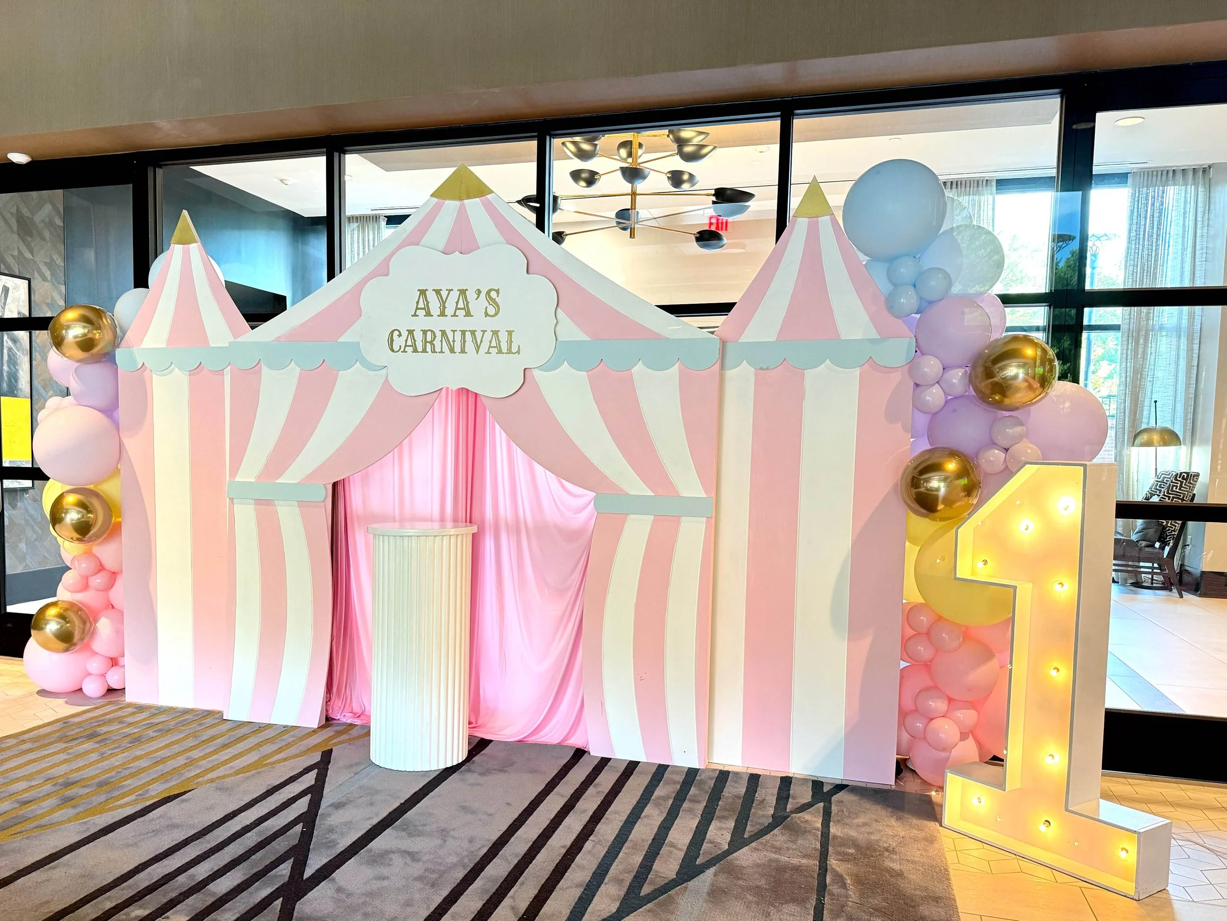 Pink and white carnival tent backdrop with balloons and a large light-up number one for a child's birthday celebration.