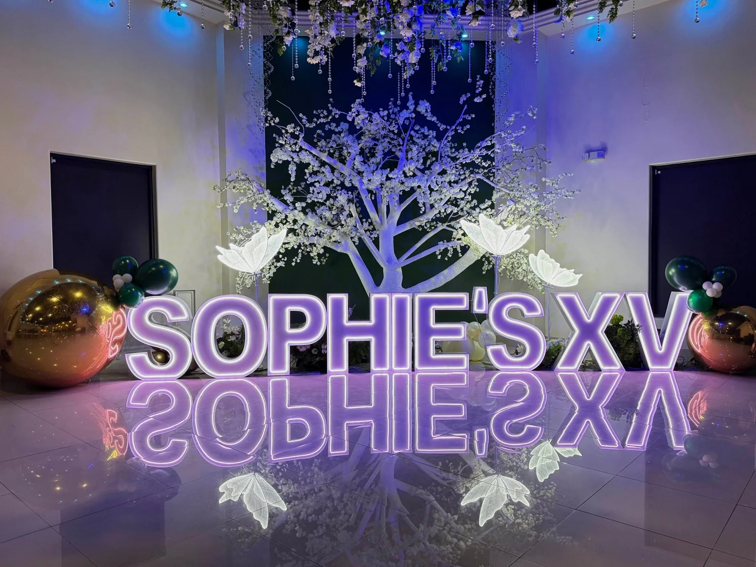 Quinceañera setup with lit purple marquee letters, a white blossom tree, glowing LED butterflies, gold mirrored spheres and a reflective white dance floor