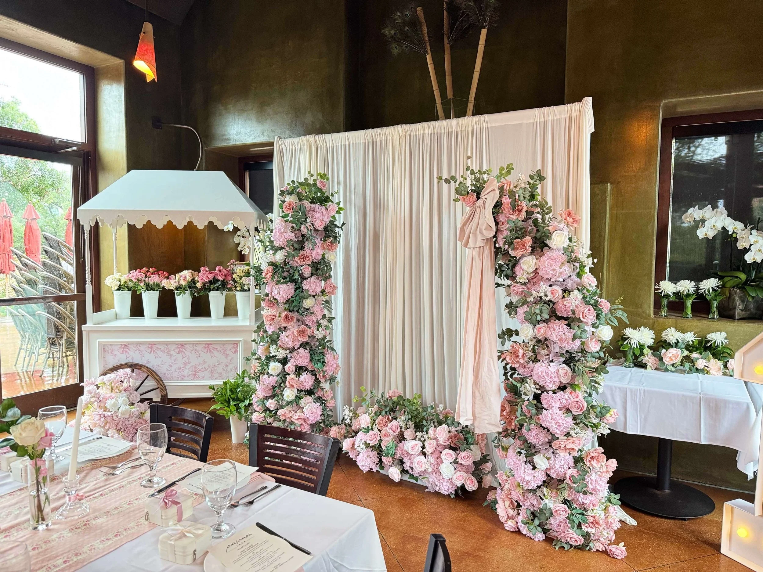 A decorated wedding backdrop with a white curtain, surrounded by pink and white flowers, in an indoor venue with windows showing outdoor patio chairs and umbrellas.