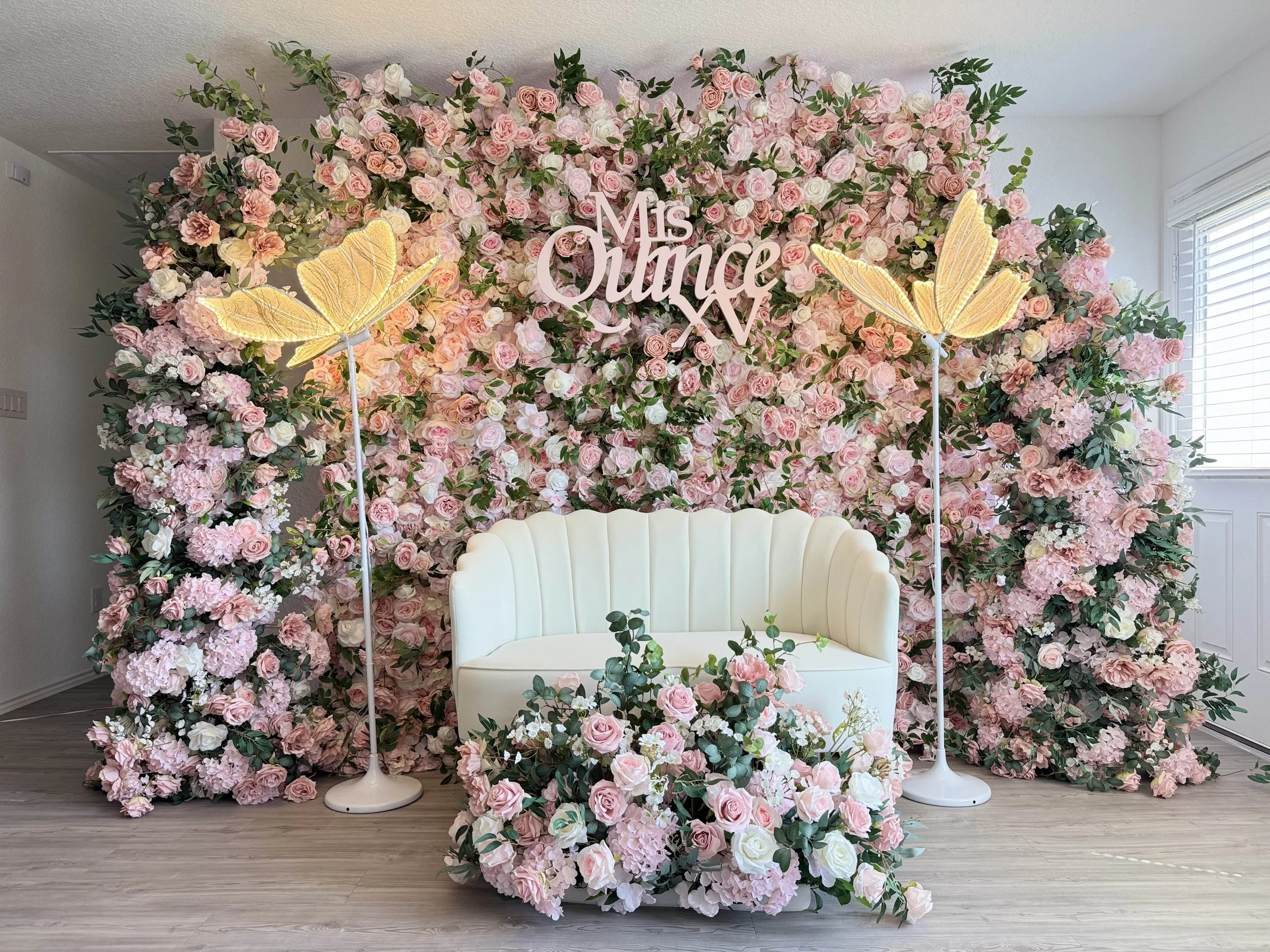 A floral backdrop with pink and white roses, green foliage, and pink lettering that reads 'Ms. Queen'. A white venue seat with a matching floral arrangement in front, flanked by tall lamp posts with yellow butterfly-shaped lights, is set against a light-colored wall and adjacent window.