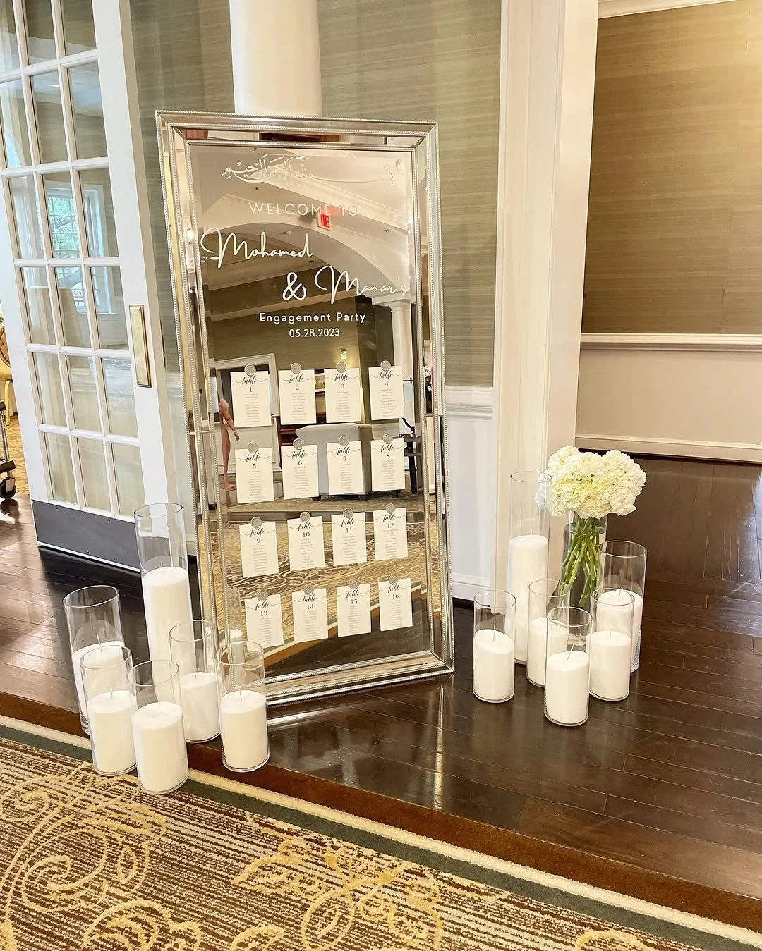 Mirror with seating chart for Mahmoud and Mona's engagement party on 05.28.2023, surrounded by white candles and flowers.