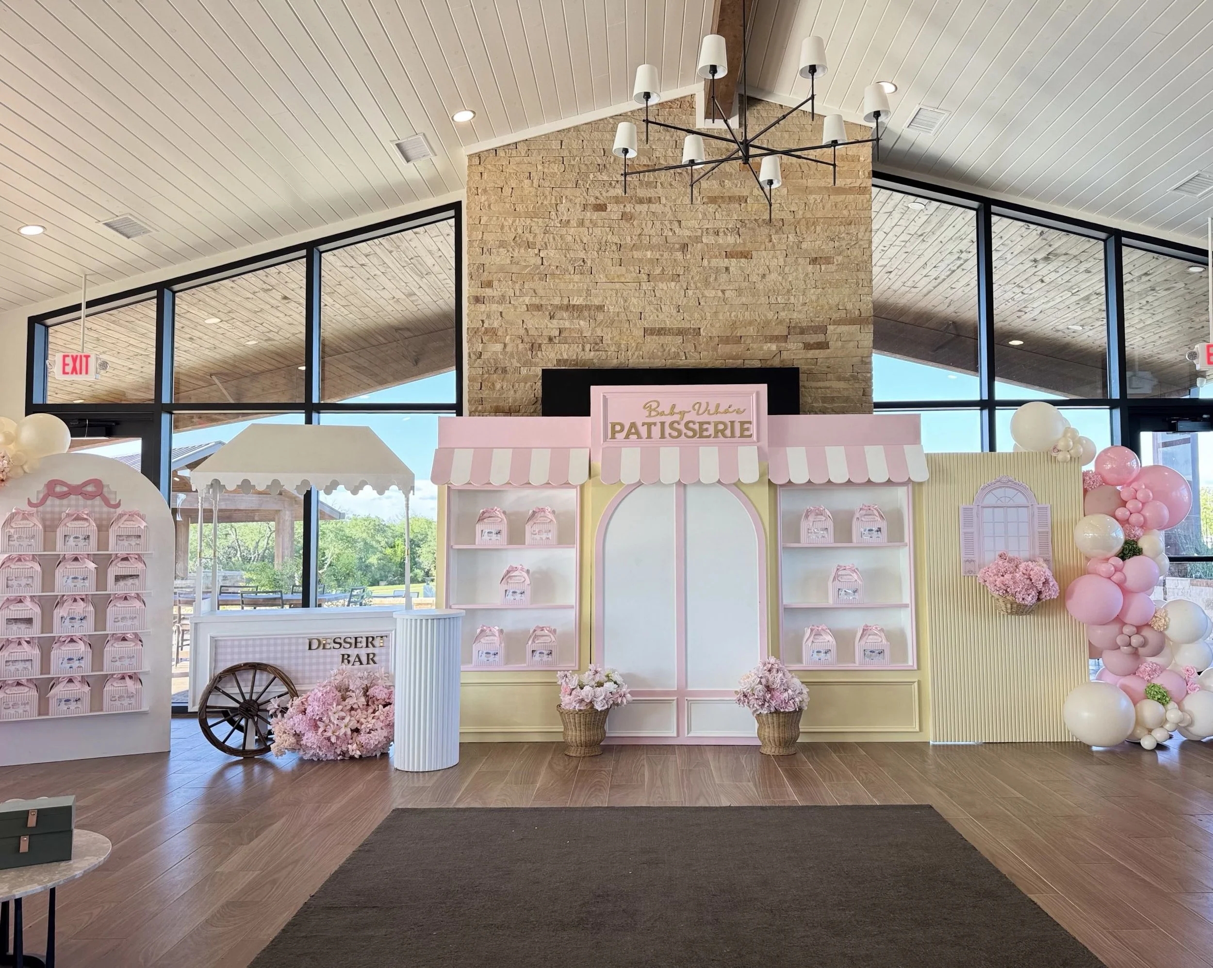 Decorative baby shower setup with pastel pink and white colors, balloons, flowers, and a sign that says "Baby Violet Patisserie" inside a venue with large windows and wood flooring.