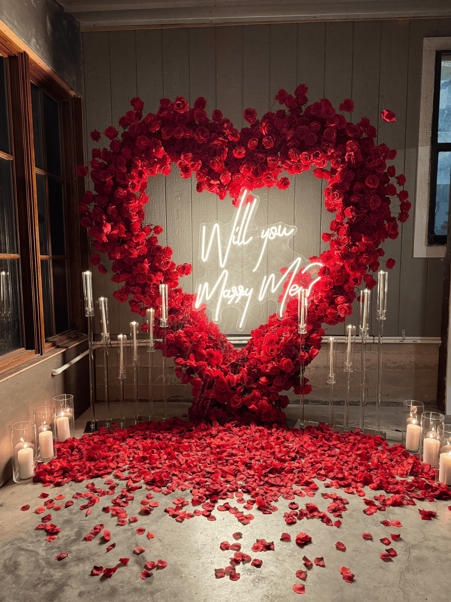 Red rose heart-shaped backdrop with a neon sign saying 'Will you Marry Me,' surrounded by candlelit glasses and scattered rose petals on the floor.