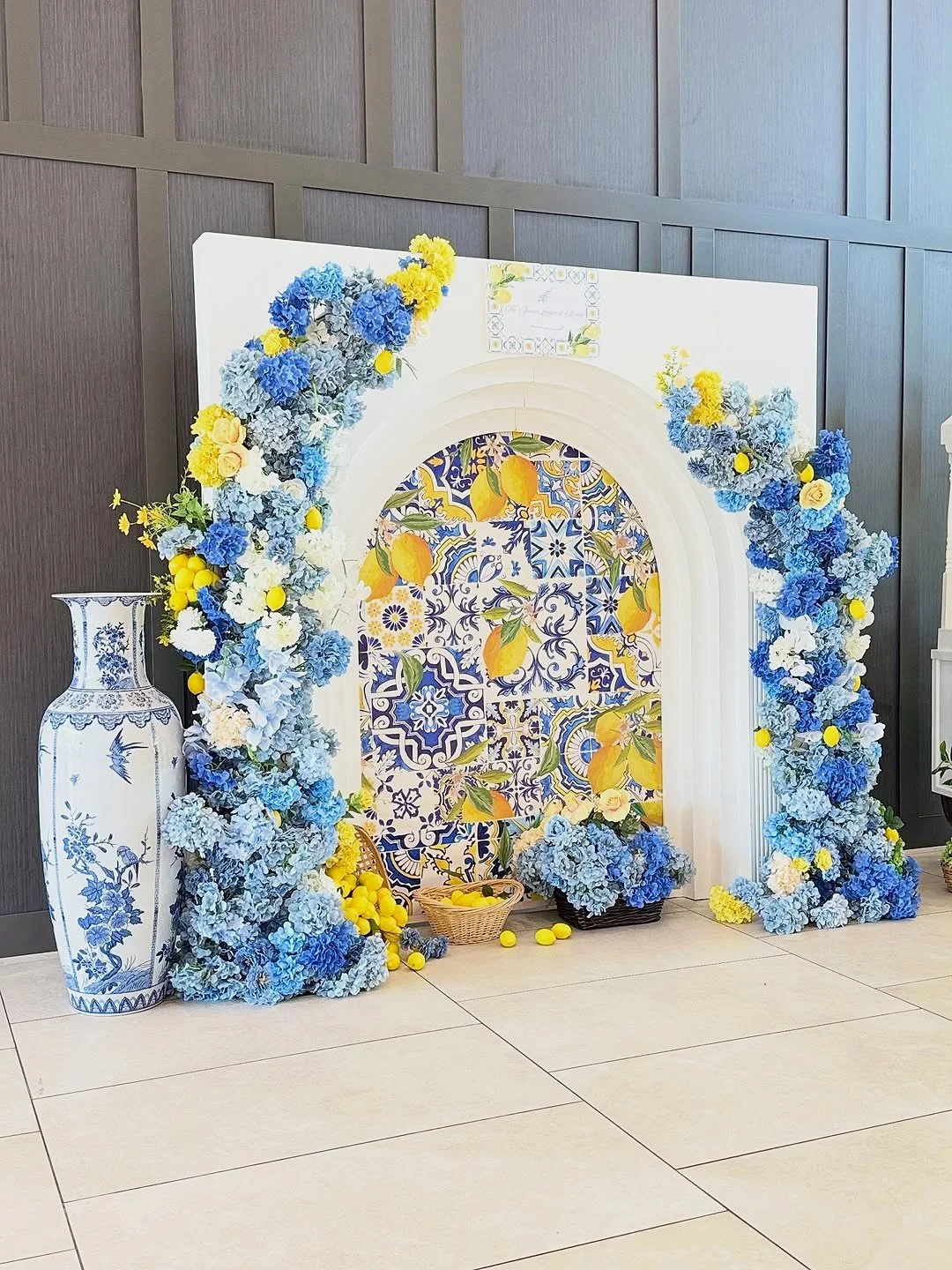 Lemon themed arch backdrop with Italian tile print, blue and yellow hydrangea floral garlands, baskets of lemons and a tall blue and white vase