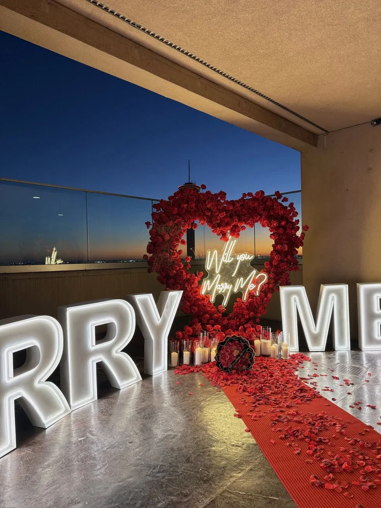 Rooftop proposal setup with large lit MARRY ME letters, a red rose heart arch with a neon sign, pillar candles, rose petals and a red carpet at dusk