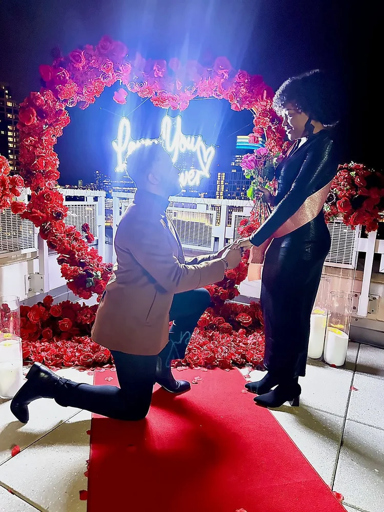 Man kneeling on a red carpet proposing under a red rose heart arch with a neon sign and candles at Mia Penthouse, the rooftop space at The Watermark Hotel in Tysons Corner, McLean, Virginia