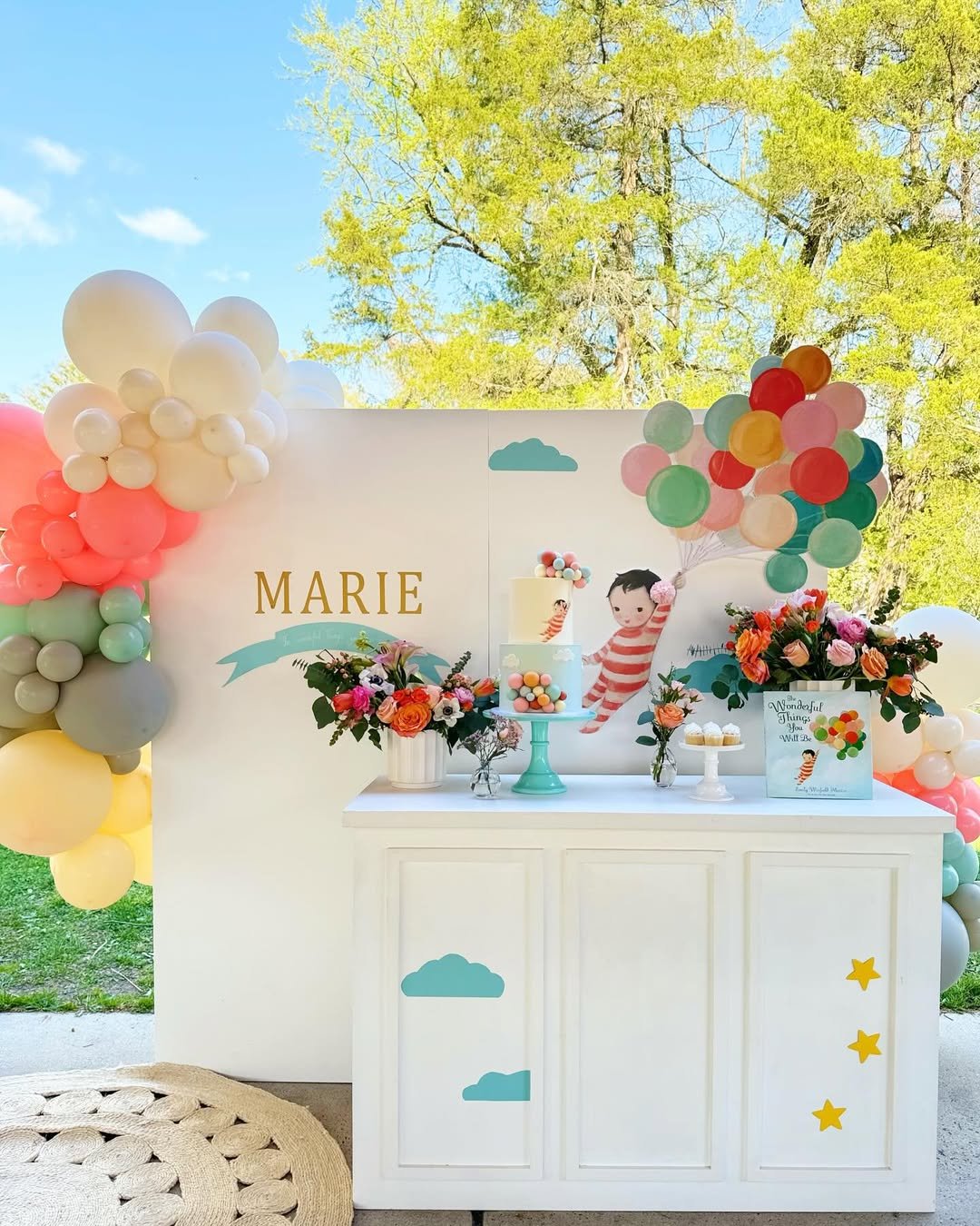 Decorated birthday party table with colorful balloons, floral arrangements, a two-tier cake, and a backdrop featuring the name Marie and a child holding balloons. Bright outdoor setting with greenery and blue sky.