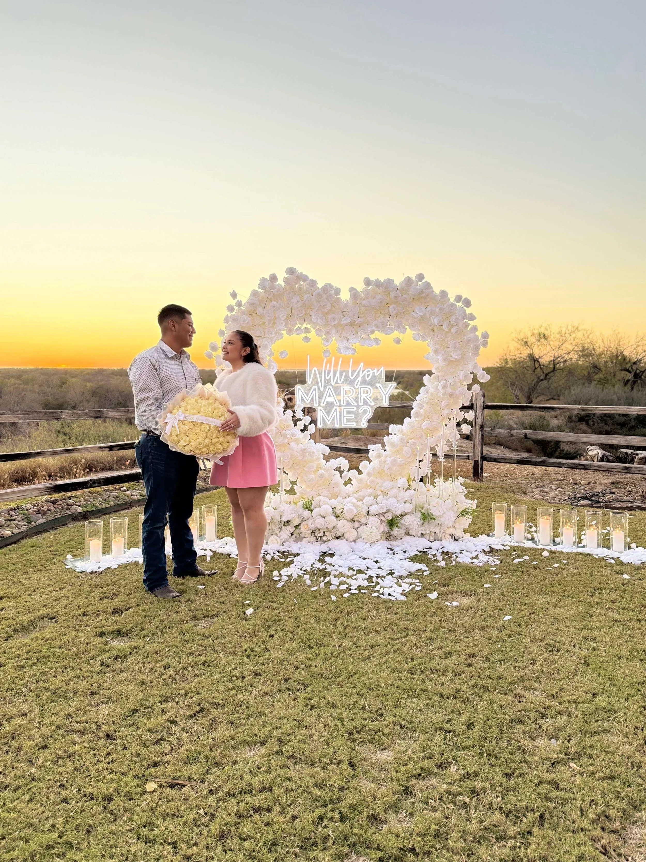 A couple standing outdoors at sunset, holding a large bouquet of yellow roses, with a floral heart-shaped backdrop and the words 'Will You MARRY ME?' illuminated in the background, surrounded by candles.