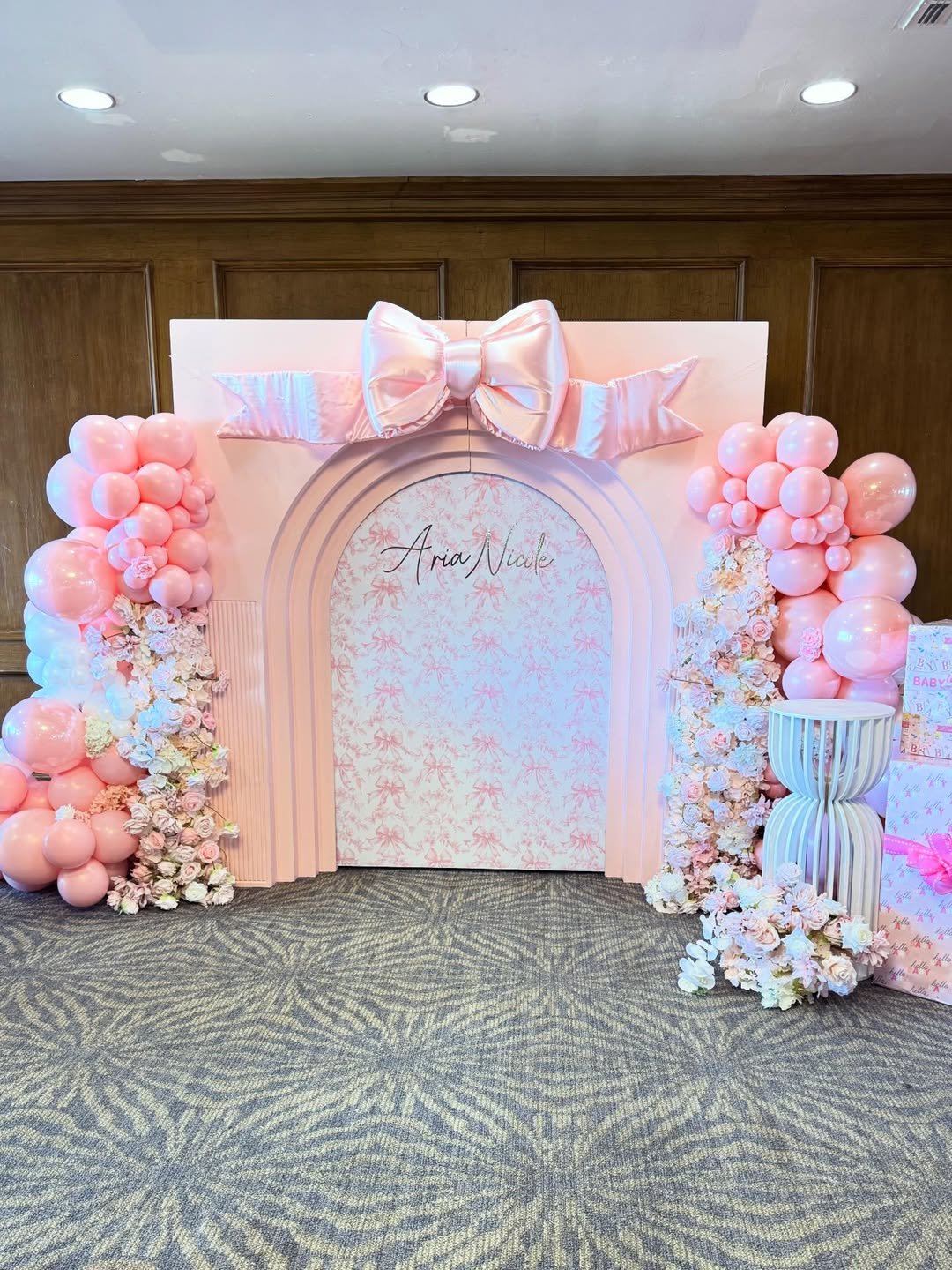 A pink-themed decorative backdrop with a large satin bow at the top center, surrounded by pink balloons and white floral arrangements on both sides, with the name 'Aria Nicole' written in the middle.