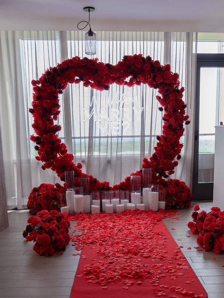 Indoor proposal setup with a red rose heart arch, Will You Marry Me neon sign, red carpet, rose petals and pillar candles in McLean, Virginia