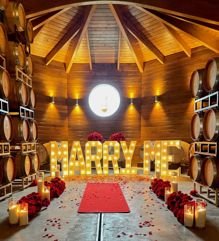 Giant lit MARRY ME marquee letters with red rose arrangements, pillar candles, a red carpet runner and rose petals in the barrel room at Greenhill Vineyards in Middleburg, Virginia