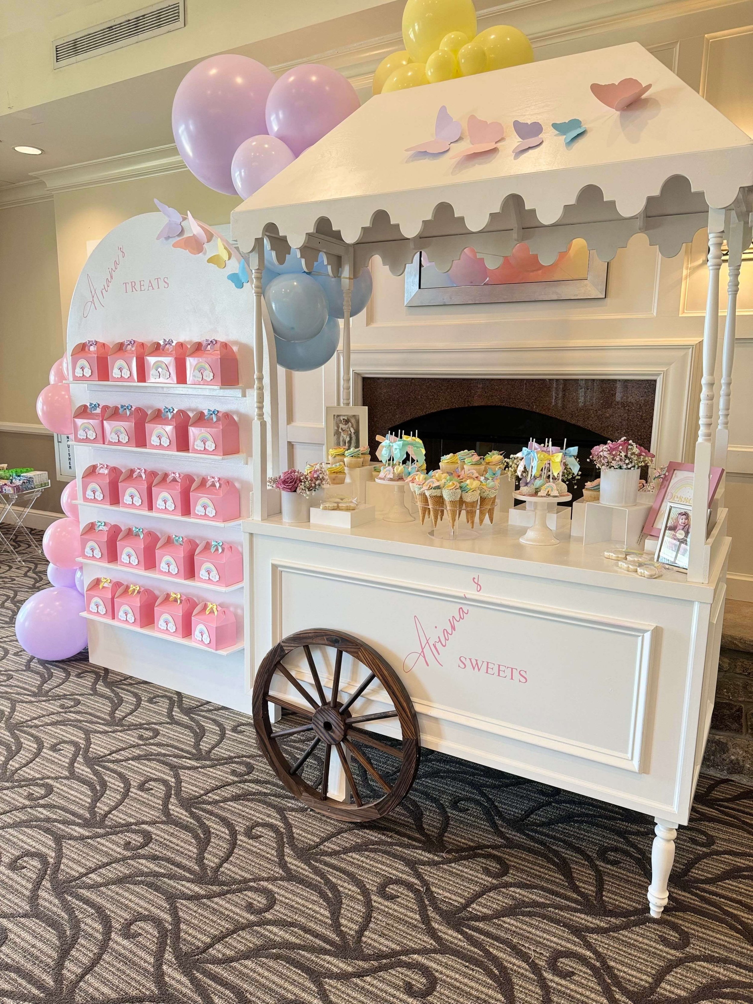 Pink and pastel-colored balloon stand alongside a white sweets cart decorated with butterflies and balloons for a celebration, featuring cupcakes and party favors.