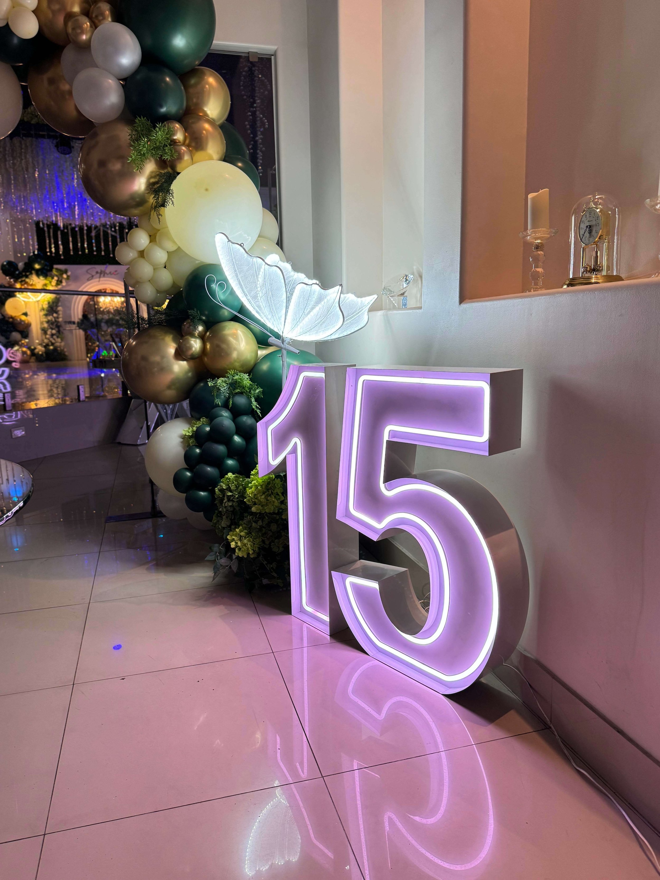 Neon number 15 sign illuminated in purple placed on a shiny floor near a wall, decorated with a large balloon arrangement in shades of green, gold, silver, and white, with a decorative butterfly and greenery, at a celebration or party venue.