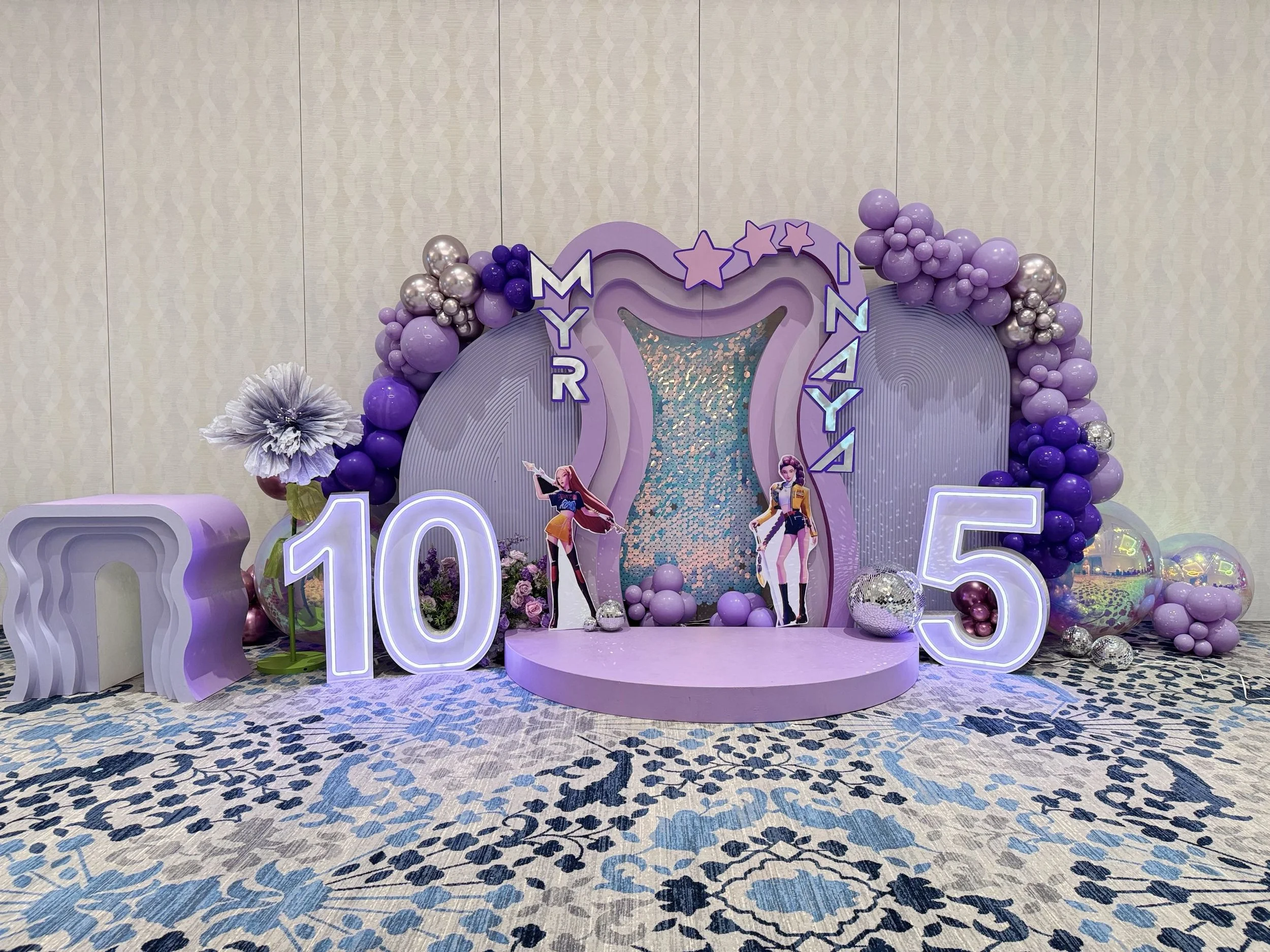 A decorated party backdrop with purple and silver balloons, Disney princess cutouts, and large illuminated numbers '10' and '05' on a patterned blue and gray carpet.