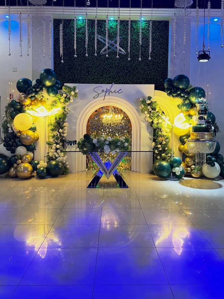 Quinceañera sweetheart setup with a white arch backdrop and custom name sign, green and gold balloon garlands, white florals, glowing LED butterflies, a mirrored table and a crystal cake stand