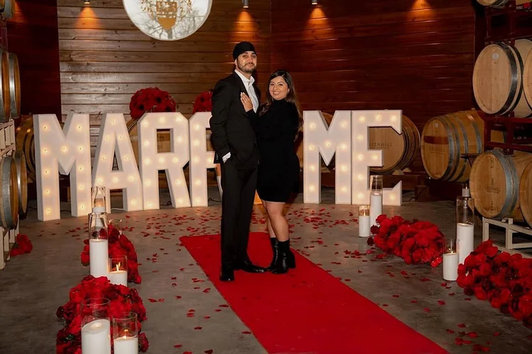 Couple celebrating their engagement in front of lit MARRY ME marquee letters on a red carpet lined with red roses and candles at Greenhill Vineyards in Middleburg, Virginia