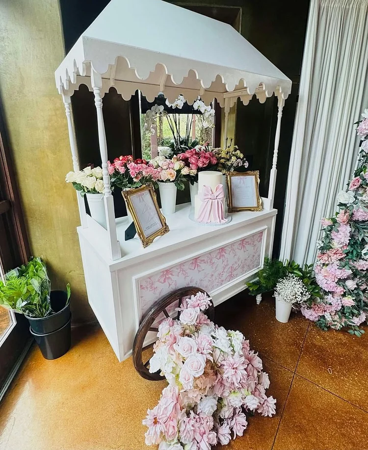 White flower cart with a scalloped roof, buckets of roses, a pink bow cake, gold framed signs and a pink bow print panel, with a loose floral arrangement on the floor