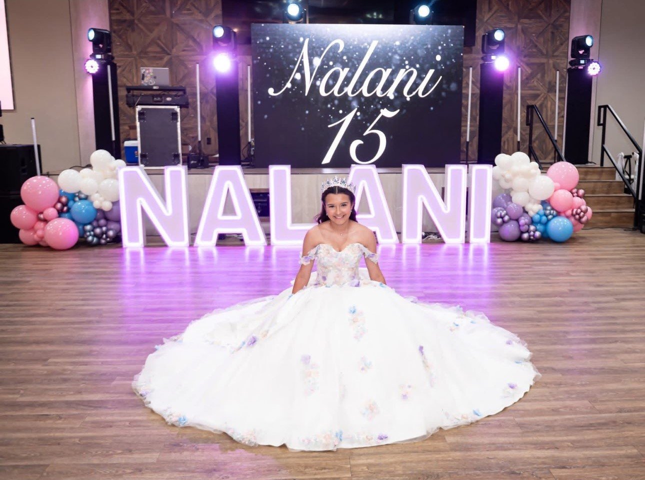 Young girl in a white floral dress with off-shoulder sleeves and a tiara, sitting on the floor with a big smile at her birthday celebration. Behind her, large illuminated letters spell 'NALANI' on the stage, which is decorated with colorful balloons on each side and a large LED screen displaying 'Nalani 15' with a starry background.