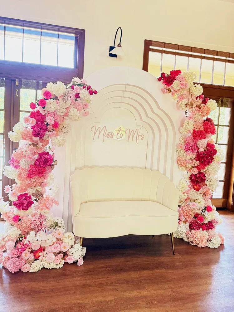 Bridal shower backdrop with a tiered arch, pink and white floral arches, Miss to Mrs neon sign and a white scalloped loveseat at Sully Station Community Association in Centreville, Virginia